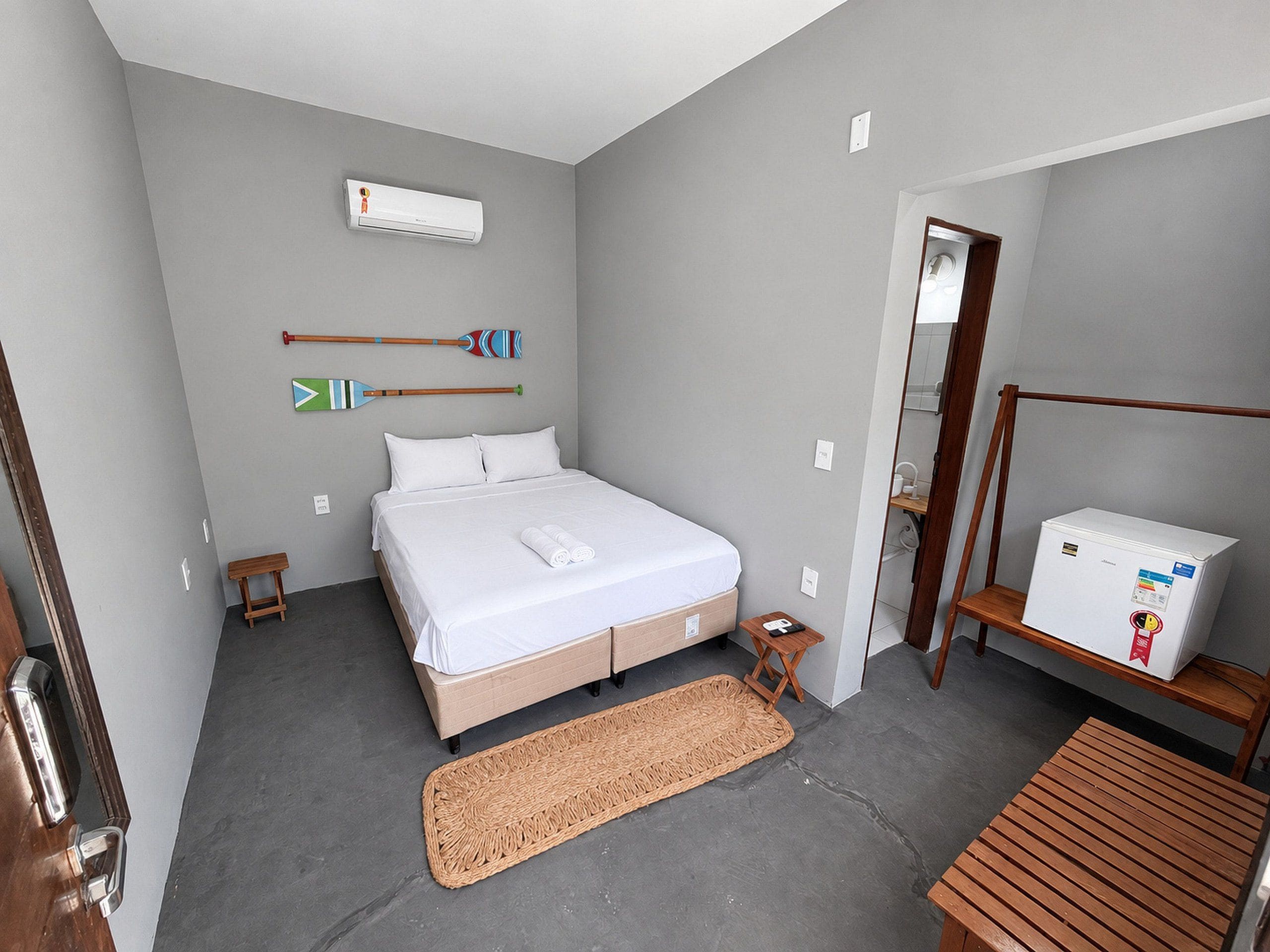 Un dormitorio con cama, una mesita de noche pequeña, aire acondicionado instalado en la pared, un banco de madera, una estera tejida y un refrigerador.
