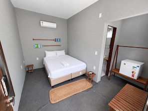 A bedroom with a bed, a small bedside table, a wall-mounted air conditioner, a wooden bench, a woven mat, and a refrigerator. - Hosts BR - Wind Suites (Barra Grande)