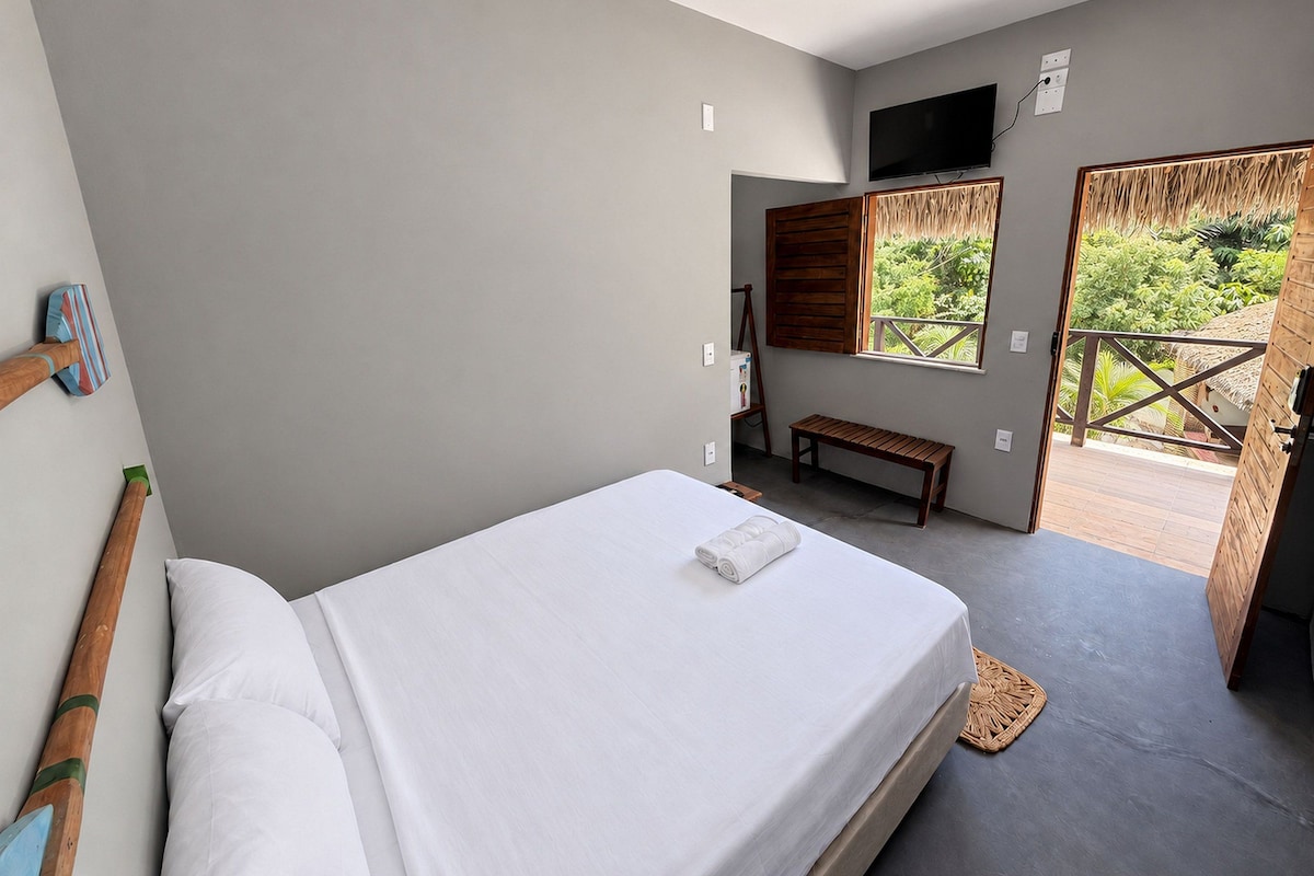 Quarto com cama, um banquinho, televisão e vista para área verde.