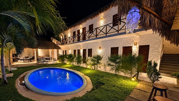 Uma área de piscina com telhado de palha, um edifício branco com venezianas de madeira e assentos ao ar livre.