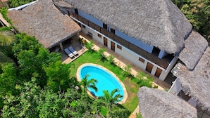 Um edifício com telhado de palha e piscina, rodeado por vegetação.