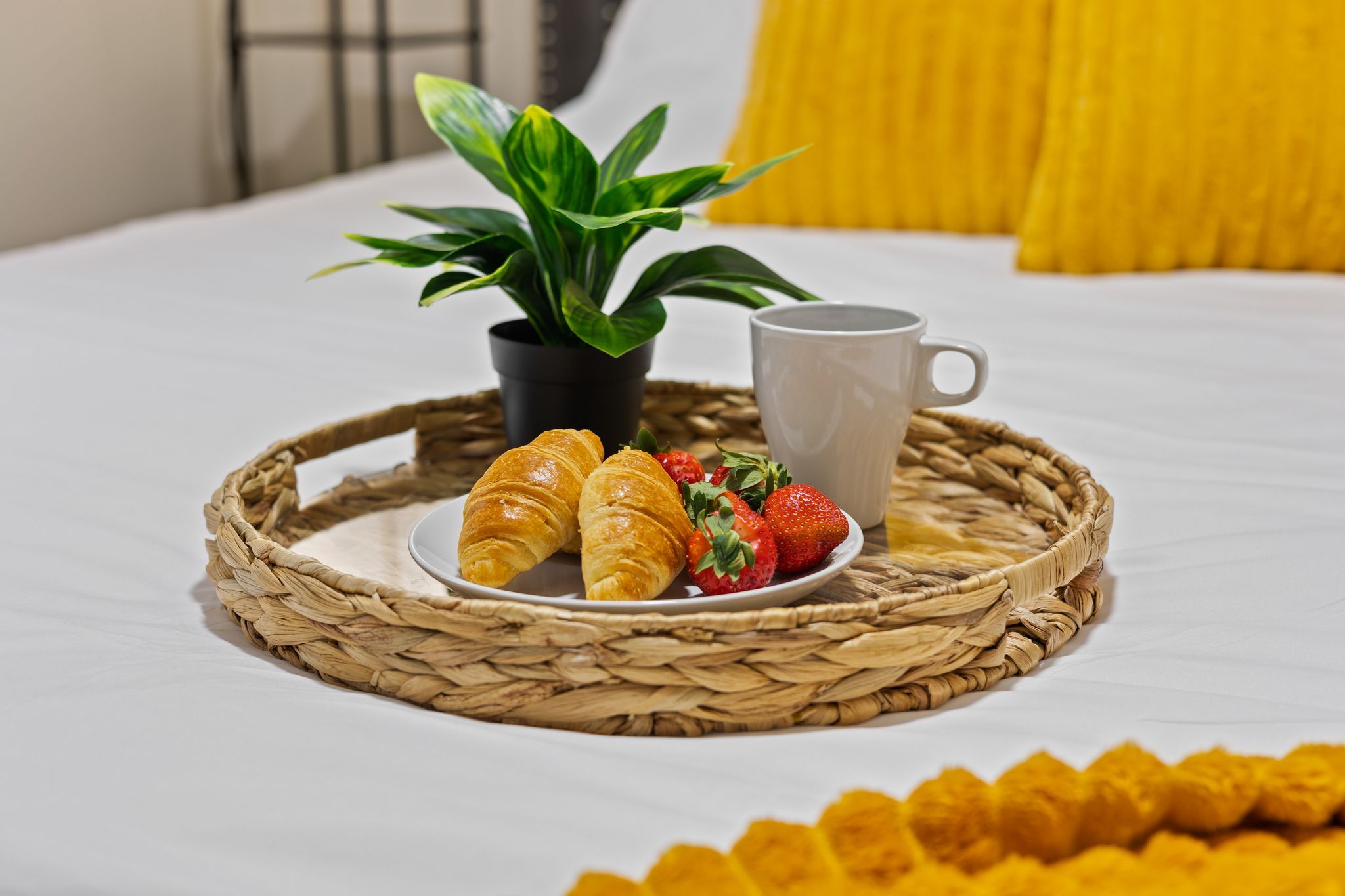 A breakfast tray with croissants, strawberries, and a mug on a bed with a white bedspread and a yellow pillow.