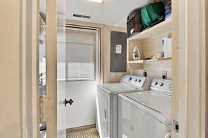 A laundry room with two washing machines, a dryer, a shelf with cleaning supplies, and a door with a window. - South Forest Beach Villa Walk to Beach and Coligny (Hilton Head Island)