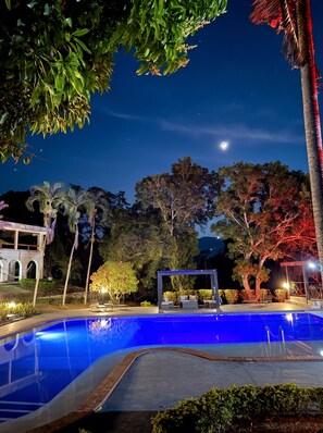 Zona de piscina nocturna con cielo despejado, una luna creciente y árboles circundantes.