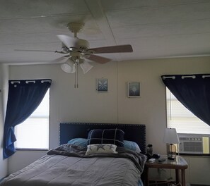 Ein Schlafzimmer mit einem Bett, einem Deckenventilator, einem Nachttisch und zwei mit Vorhängen versehenen Fenstern.