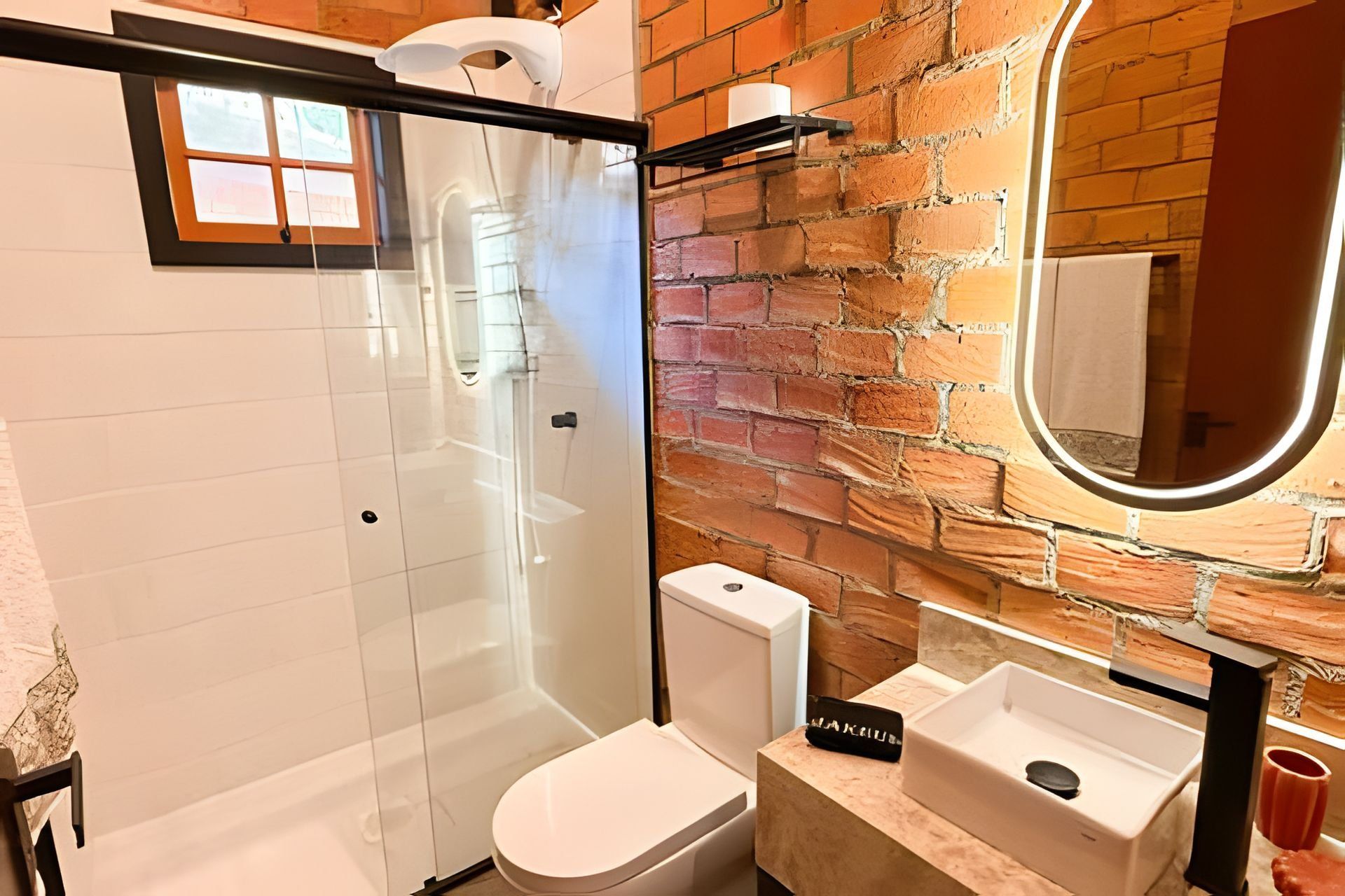 A bathroom with a glass-enclosed shower, a white toilet, a sink with a mirror, and a brick wall.