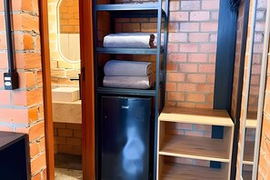 A compact room with a brick wall, a small black refrigerator, and a wooden shelf unit with folded towels. - ASA Quinta da Esmeralda Bar Pool Cabins (Itajai)