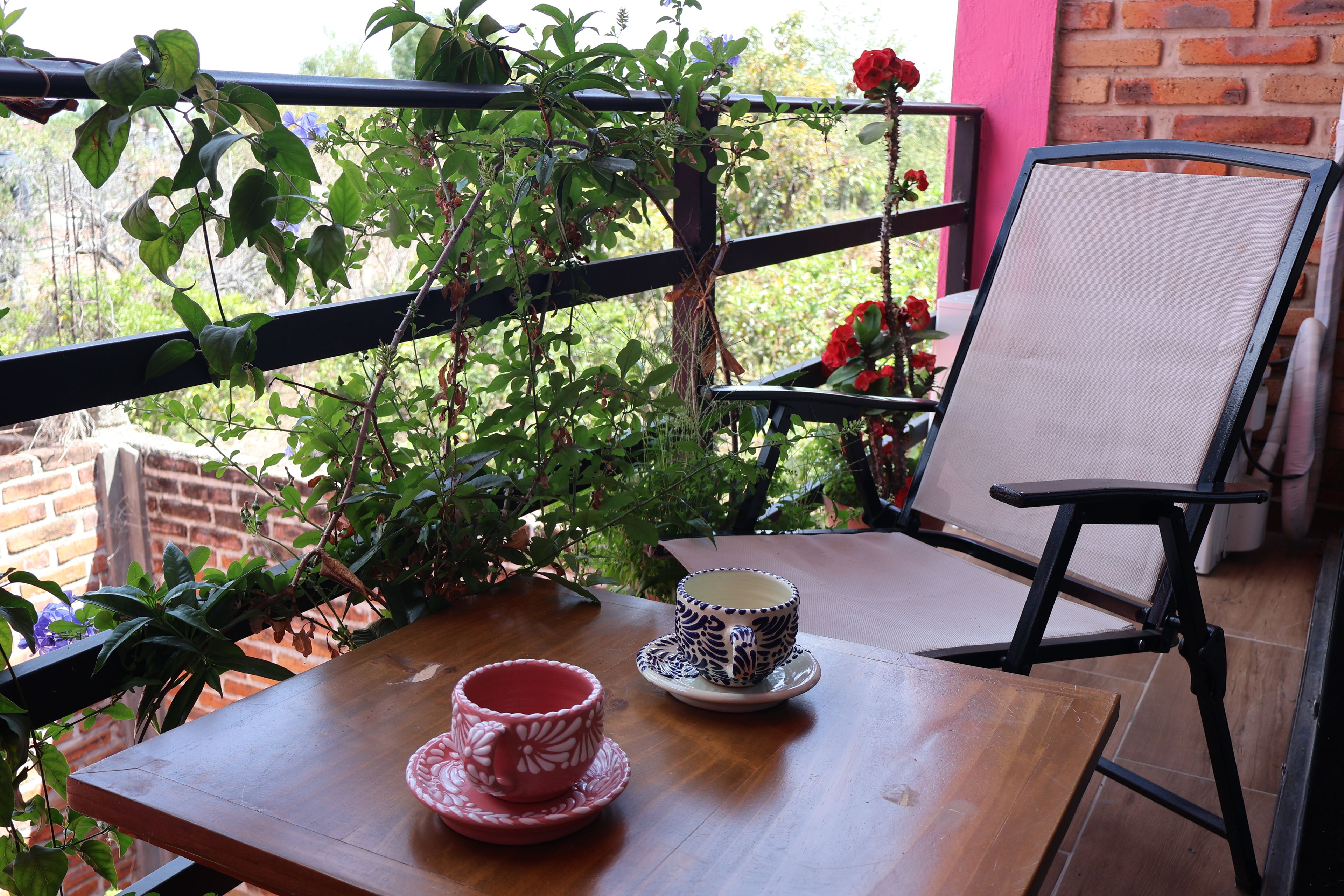 Een houten tafel met twee bekers en een stoel op een balkon met planten en een bakstenen muur.
