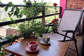 A wooden table with two cups and a chair on a balcony with plants and a brick wall.