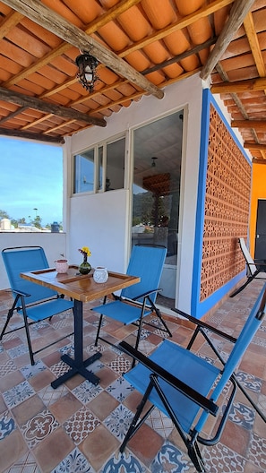 A covered patio with a table and blue chairs, a view of the outside, and a patterned floor.