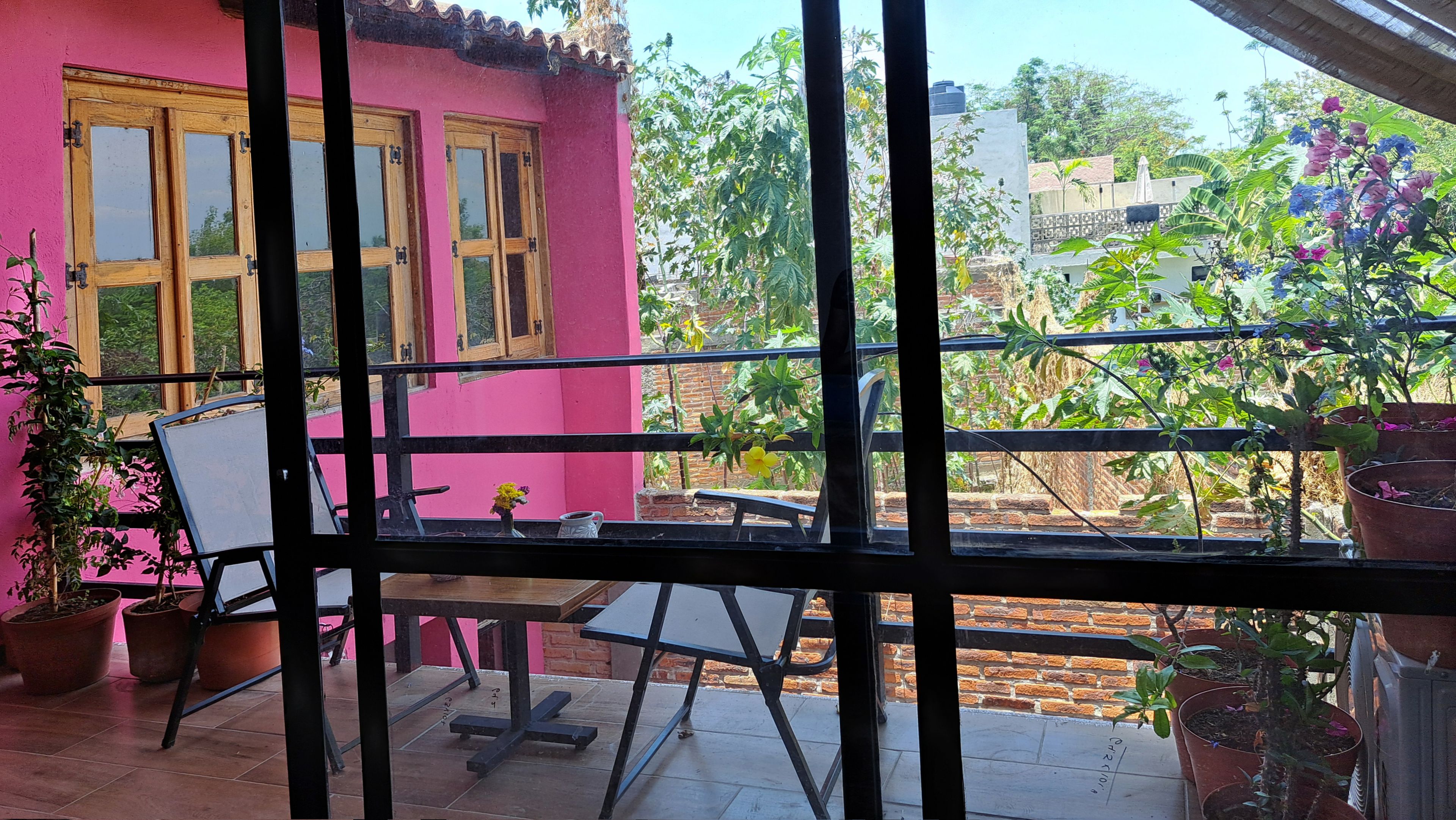 Un balcón con mesa y sillas, plantas en macetas y un edificio rosa con ventanas.