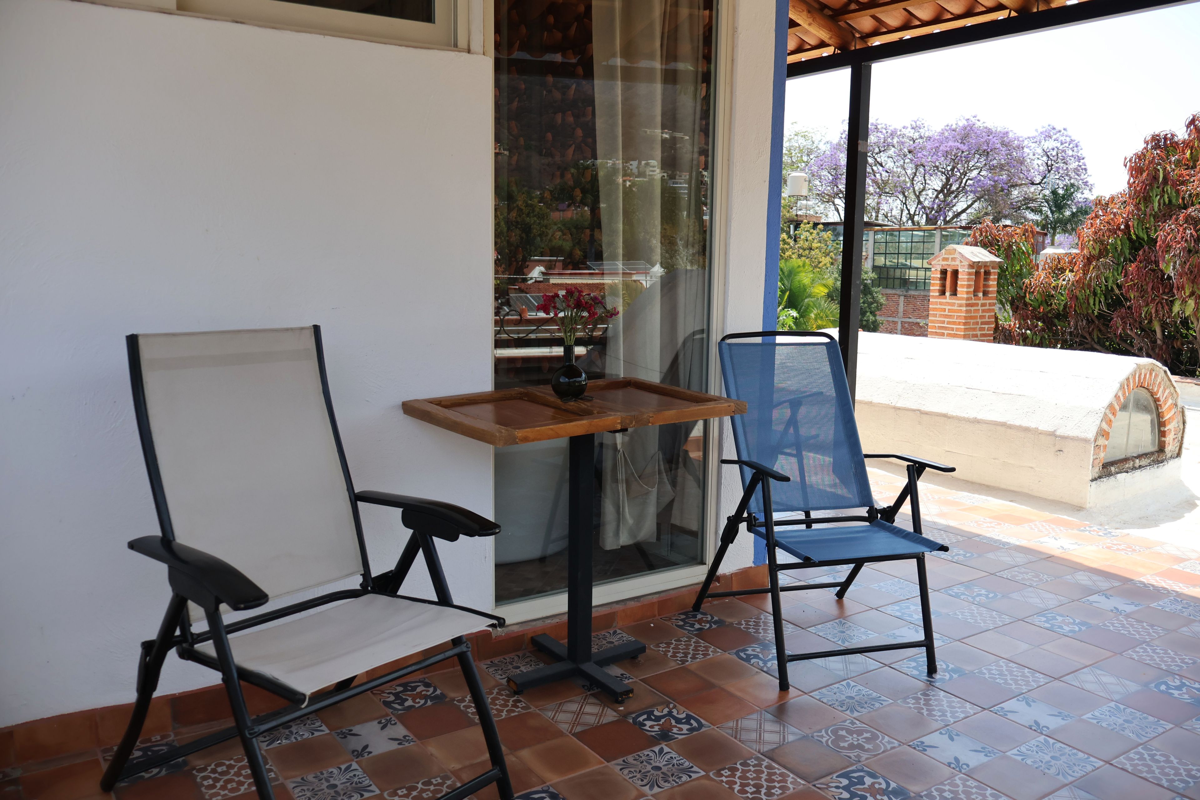 Un espacio de patio con dos sillas, una mesita y vista a un jardín a través de un ventanal.