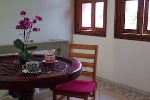 A round wooden table with a vase of pink flowers, two patterned cups, and a wooden chair with a pink cushion.