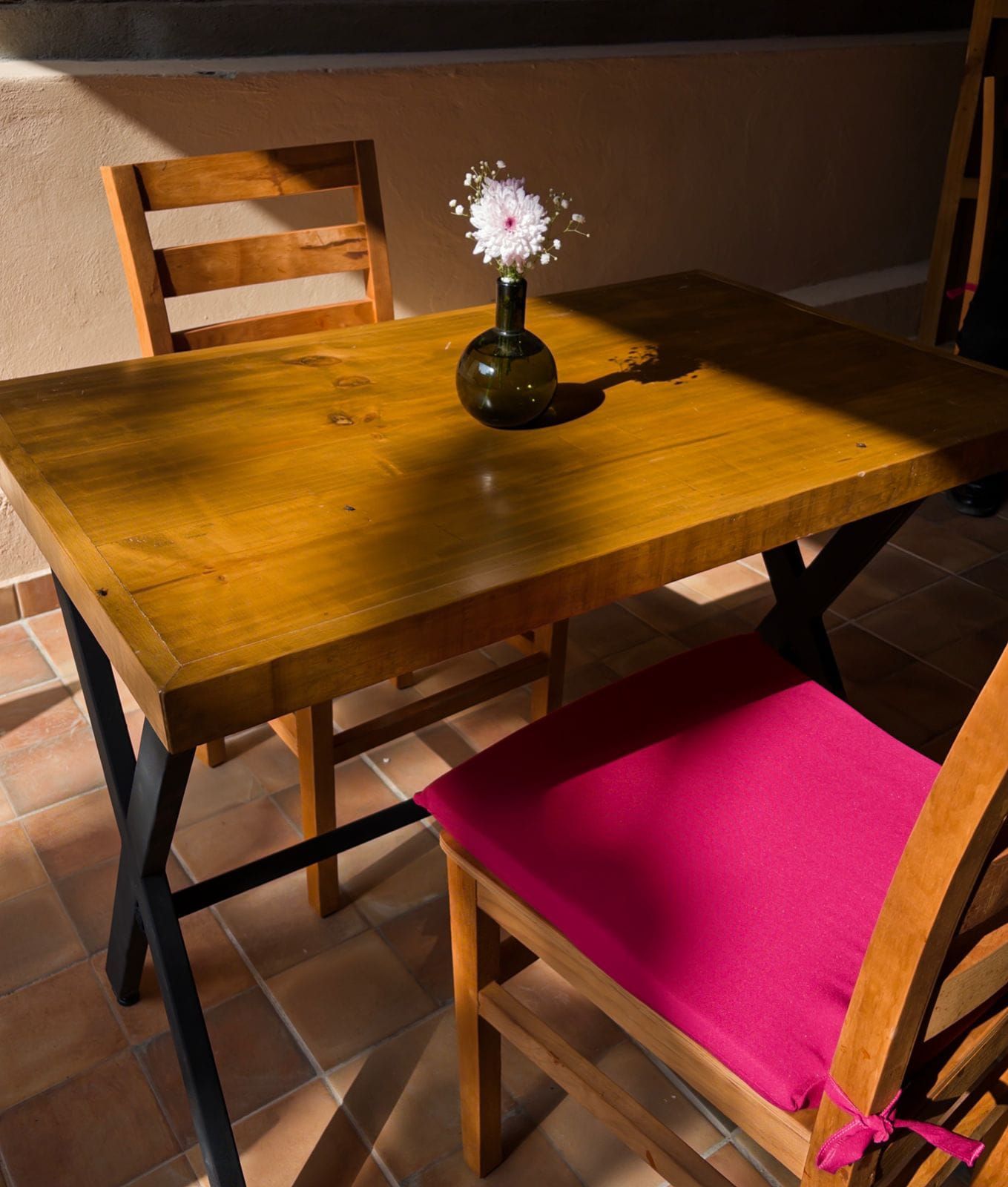 Uma mesa de madeira com um vaso de flores, uma cadeira com almofada rosa e um chão ladrilhado.
