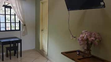 Um quarto com piso ladrilhado, uma escrivaninha com cadeiras, uma televisão fixada na parede e uma mesinha com um vaso de flores.