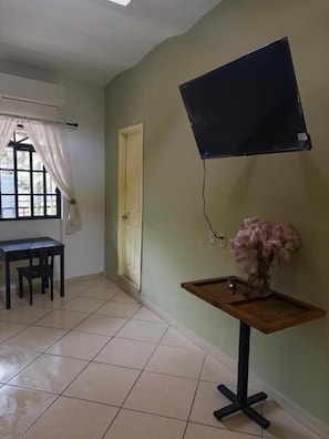 A room with a tiled floor, a desk with chairs, a television mounted on the wall, and a small table with a vase of flowers.