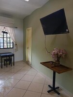 Una habitación con piso de baldosas, un escritorio con sillas, un televisor montado en la pared y una pequeña mesa con un florero.
