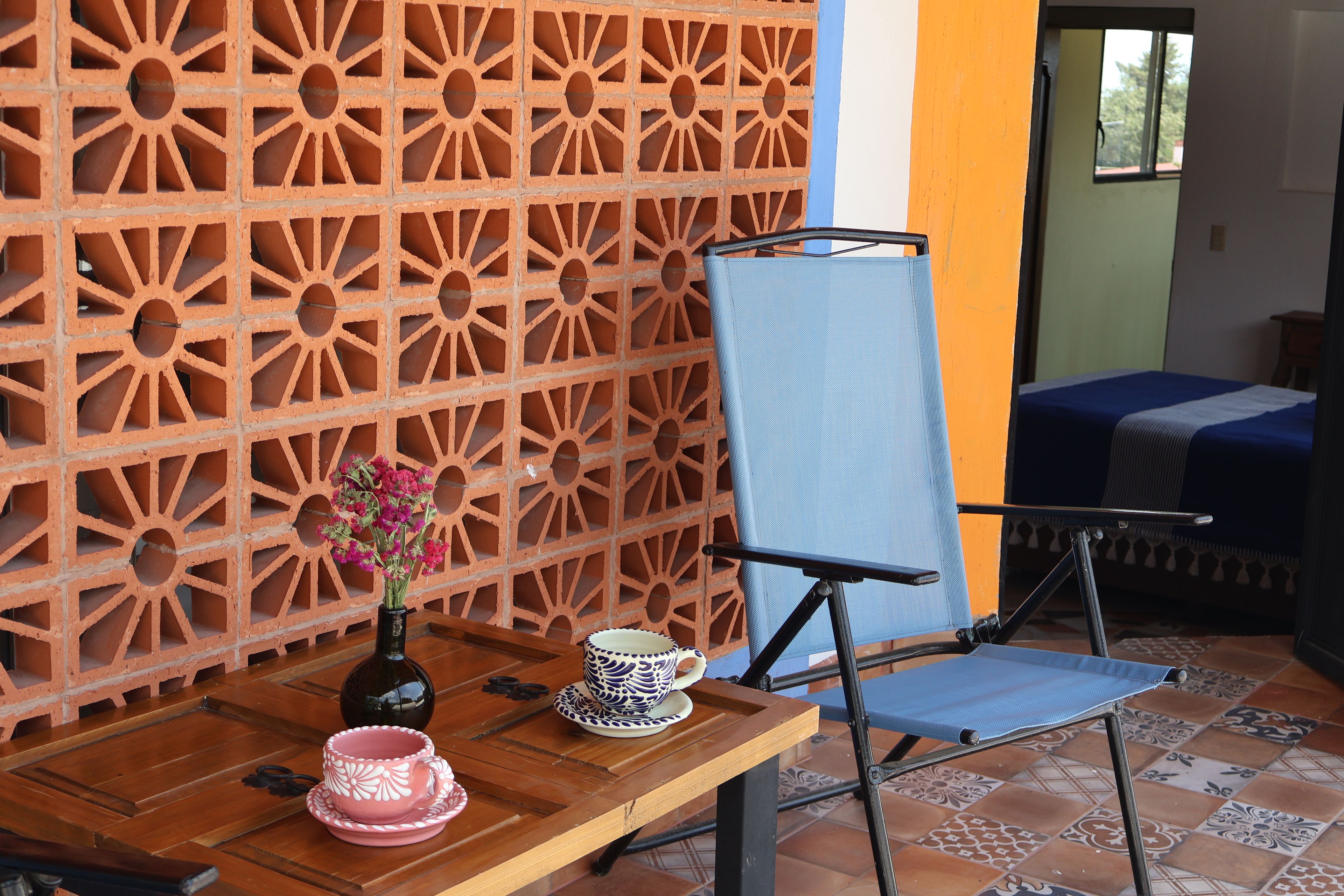 Una habitación con pared de ladrillo con diseño, una silla plegable azul, una mesa de madera con una taza y platillo rosas, y una cama con cubrecama azul.