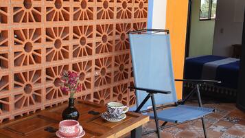 Una habitación con pared de ladrillo con diseño, una silla plegable azul, una mesa de madera con una taza y platillo rosas, y una cama con cubrecama azul.