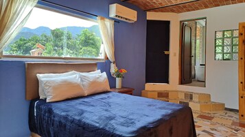 Un dormitorio con cama, ventana con cortinas, aire acondicionado instalado en la pared y vista a zonas verdes al exterior.