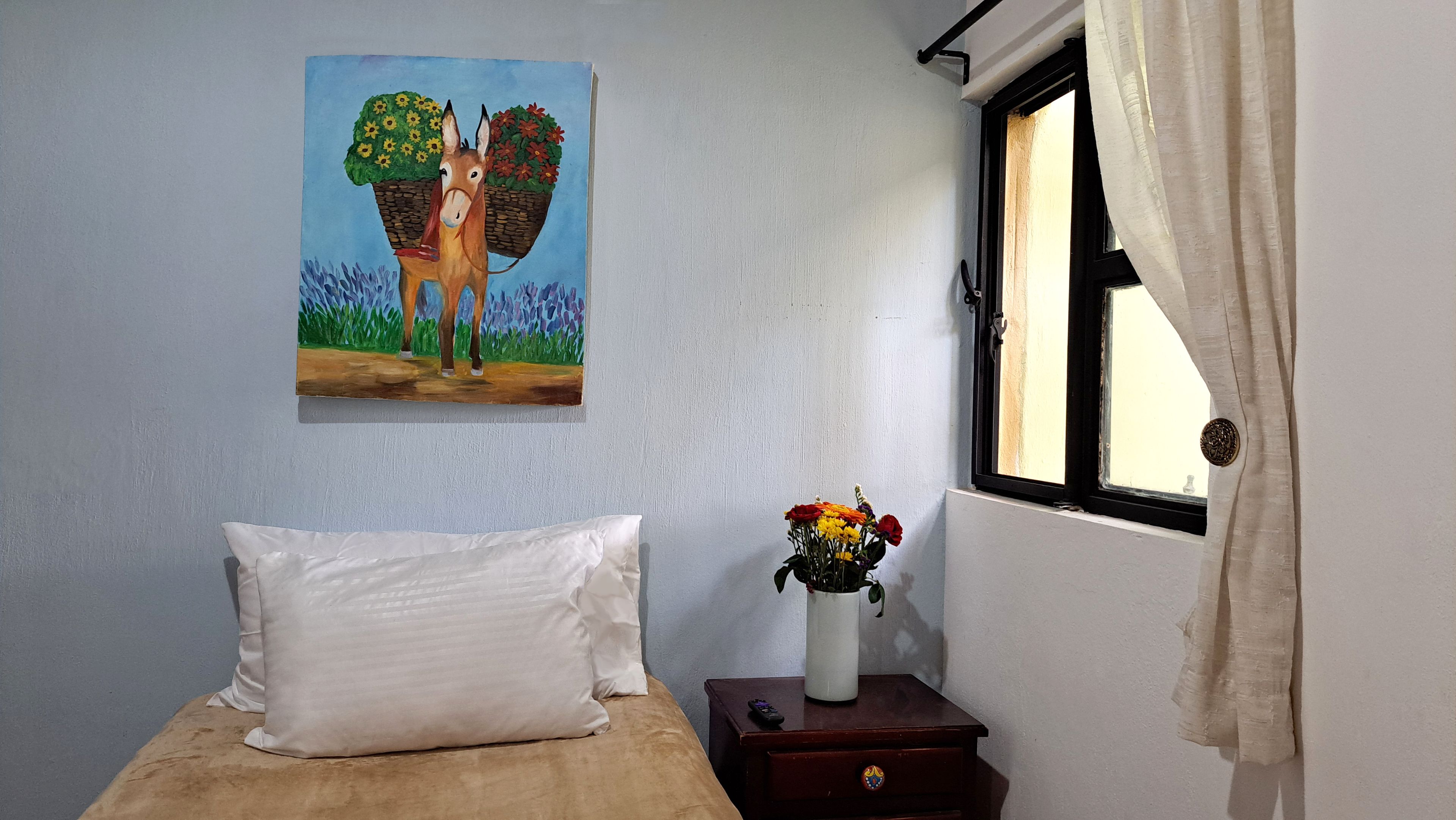 Una habitación con una cama, un cuadro de un caballo con una cesta, un florero con flores y una ventana con cortinas.