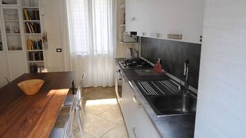 A modern kitchen with a wooden dining table, clear chairs, a built-in oven, and a window with sheer curtains.