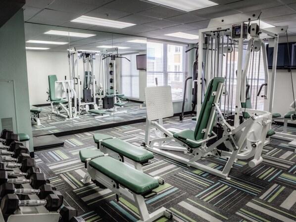 Een sportschool met diverse fitnessapparaten, een spiegelwand en een kamerbreed tapijt met een patroon.