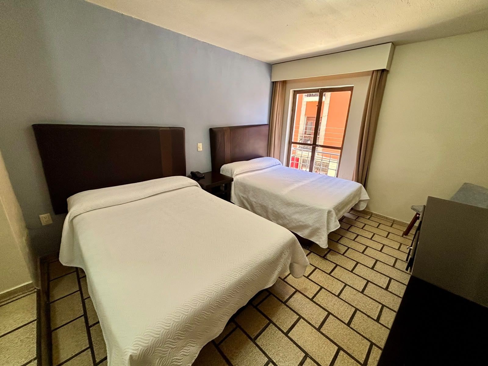 A hotel room with two beds, a small table, and a window with curtains.