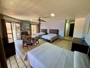 Une chambre d’hôtel avec deux lits, une table à manger avec des chaises, un ventilateur de plafond et un tableau au mur.