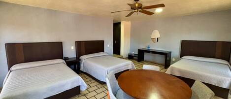 A hotel room with three single beds, a round wooden table with two chairs, a ceiling fan, and a small desk with a lamp.