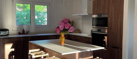 Cocina moderna con una isla central, taburetes de madera, iluminación colgante y vistas a zonas verdes.