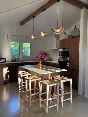 A modern kitchen with a central island, wooden bar stools, pendant lighting, and a view of greenery outside. - A cozy house with a garden, located between the ocean, the Landes region, and the Pyrenees (Angoumé)