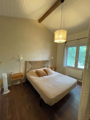 A bedroom with a bed, bedside tables, a standing heater, and a window with a view of greenery. - A cozy house with a garden, located between the ocean, the Landes region, and the Pyrenees (Angoumé)