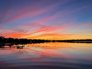 Tyyni vesialue, jonka pinnasta heijastuu auringonlaskun väriloisto. Taustalla näkyy laituri ja puita.