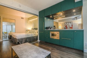 A modern hotel room with a bed, a small table, a kitchenette with a microwave, and a glass partition separating the bedroom from the living area. - Premium 1BR Condo | Balcony + Prime Location (Toronto)