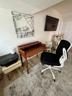 A room with a wooden desk, a black office chair, a small white table, and a wall-mounted TV.