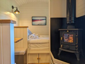 A cozy room with a bed, a wooden bench, a wall-mounted lamp, a painting, and a wood-burning stove.