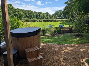 Un espace extérieur aménagé avec des sièges en bois et un jacuzzi, entouré d’un jardin clos, et offrant une vue sur un champ verdoyant et des arbres au loin.