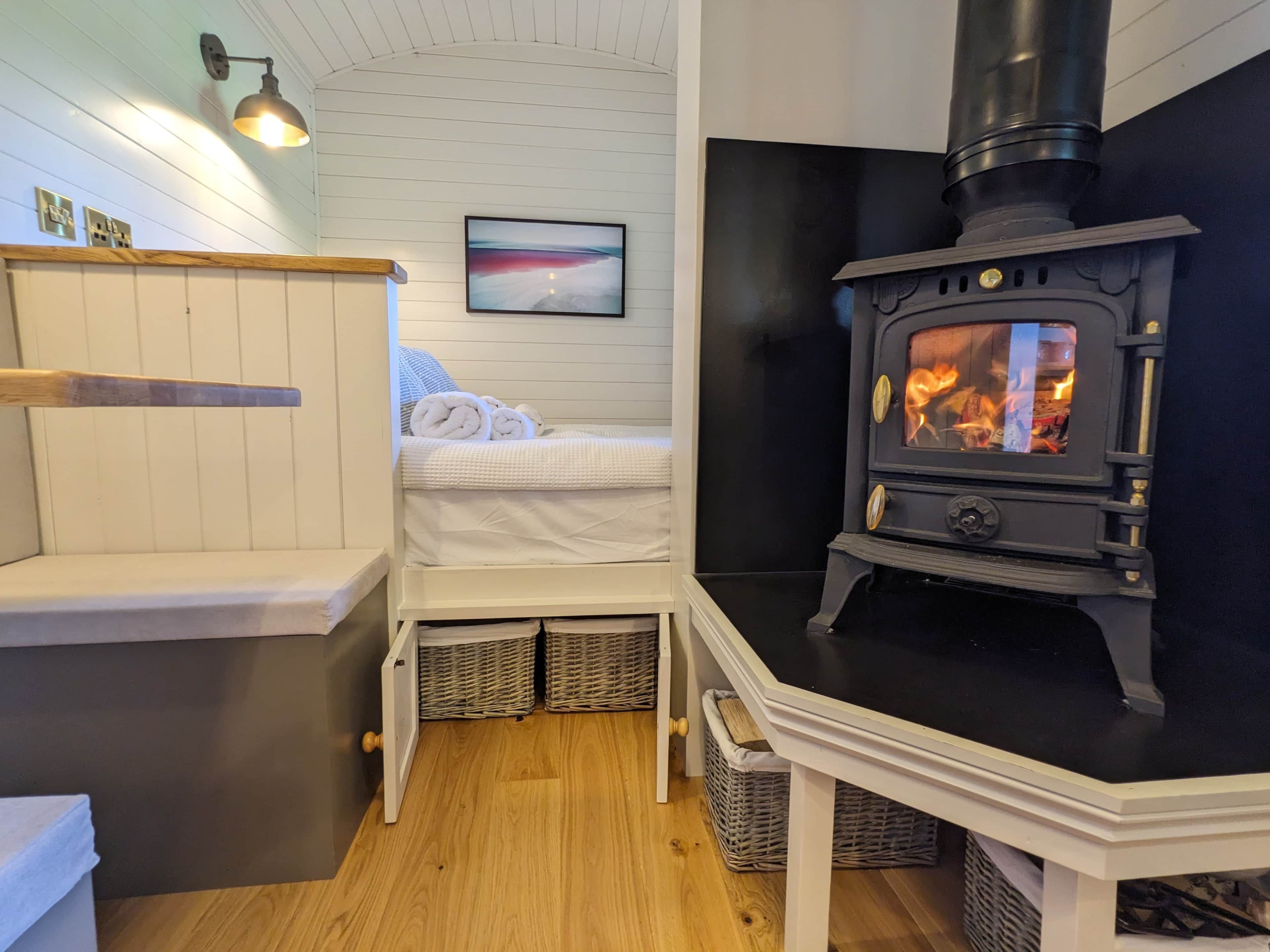 Une pièce chaleureuse avec un lit, un banc blanc, une lampe fixée au mur, un tableau et un poêle à bois.