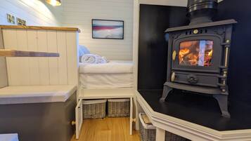Une pièce chaleureuse avec un lit, un banc blanc, une lampe fixée au mur, un tableau et un poêle à bois.