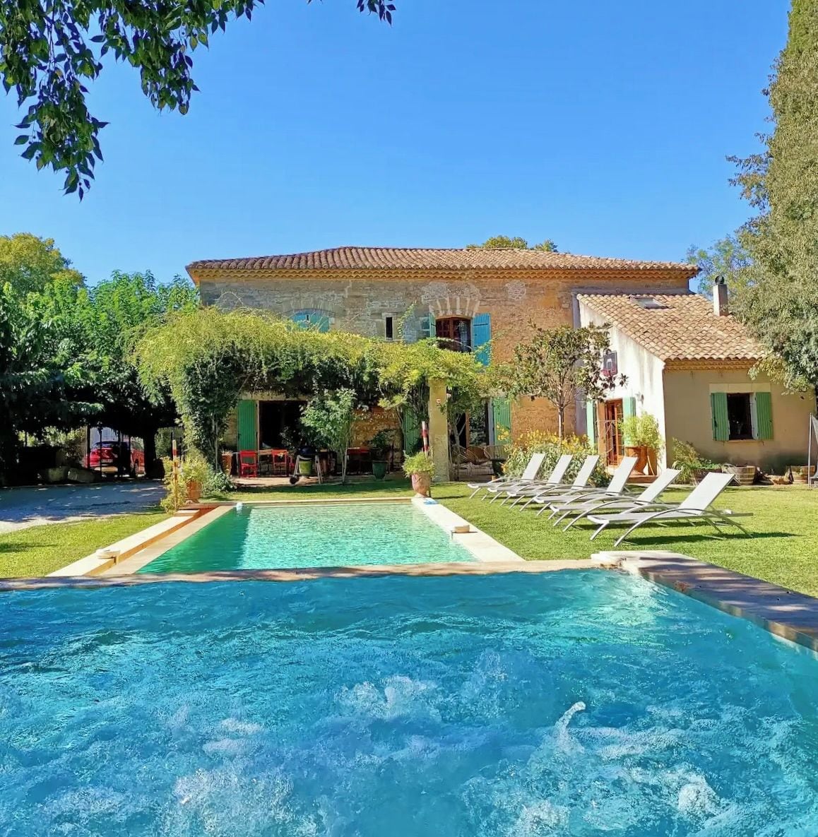 A swimming pool with lounge chairs, a stone building with a tiled roof, and greenery.