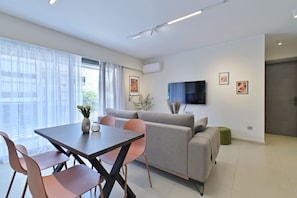 A modern living room with a sofa, a dining table with chairs, a flat-screen TV mounted on the wall, and a window with sheer curtains. - Bright And Elegant Apartment - Lotis (Dafni)