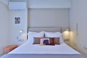 A hotel room with a bed, pillows, a bedside table, a wall-mounted air conditioner, a light fixture, and a framed artwork. - Bright And Elegant Apartment - Lotis (Dafni)