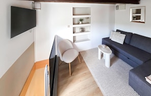 Ein modernes Wohnzimmer mit einem an der Wand befestigten Flachbildfernseher, einem grauen Sofa mit weißem Zierkissen, einem weißen Sessel und einem kleinen weißen Beistelltisch.