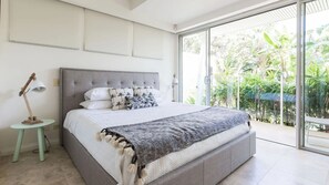 Une chambre moderne avec un grand lit, une petite table de chevet et une vue sur la verdure grâce à une porte-fenêtre coulissante.