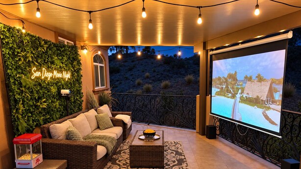 Un espace de détente extérieur accueillant, avec un canapé, une table et un écran de projection diffusant une scène de plage.