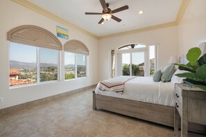 Quarto com cama, ventilador de teto e janelas amplas com vista para a cidade.