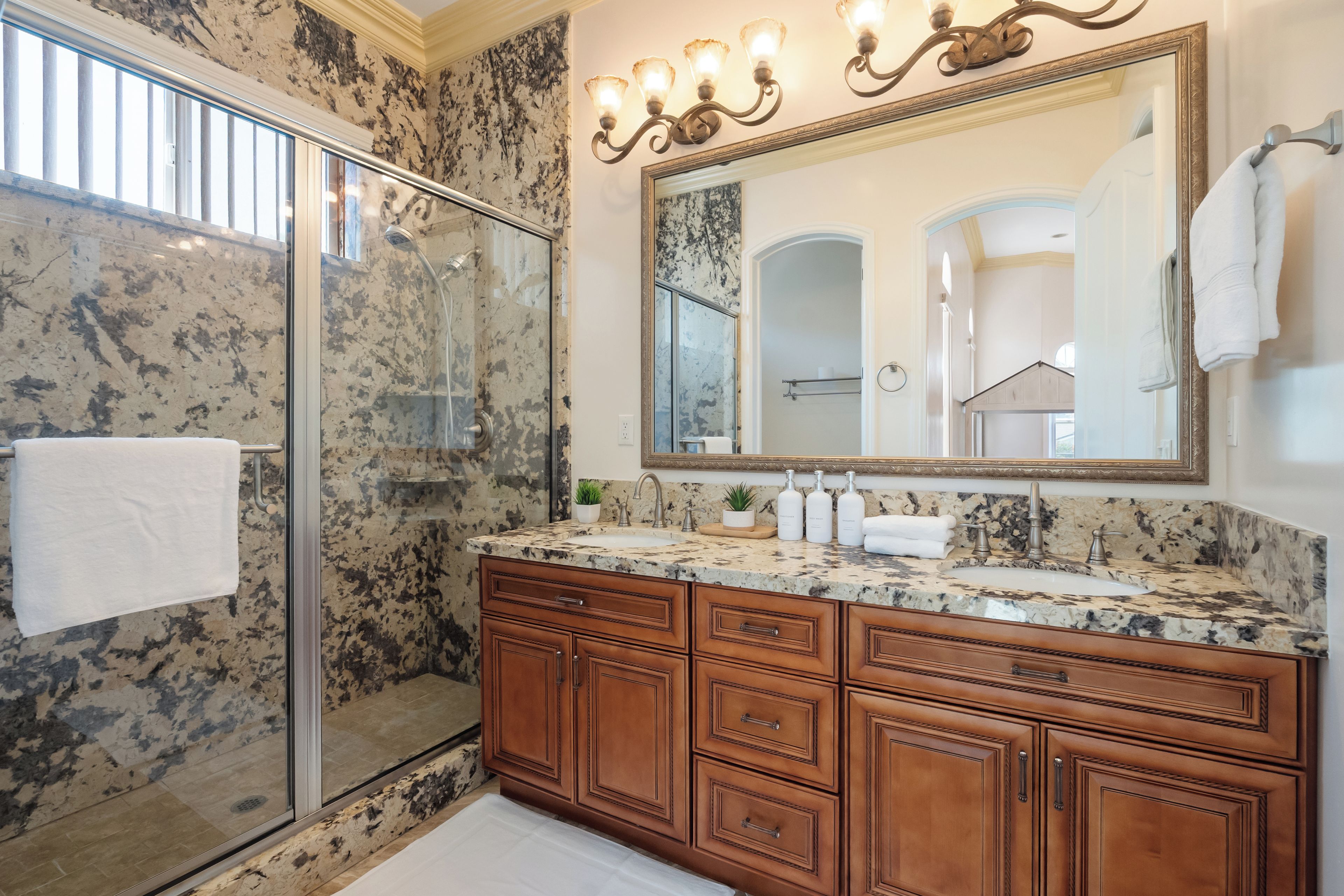 A bathroom with a glass shower enclosure, a marble countertop, wooden cabinetry, and a large mirror with wall sconces.