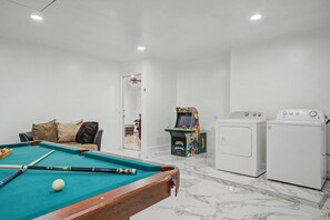 Een kamer met een pooltafel, een bank met kussens, een spelconsole en een wasmachine.