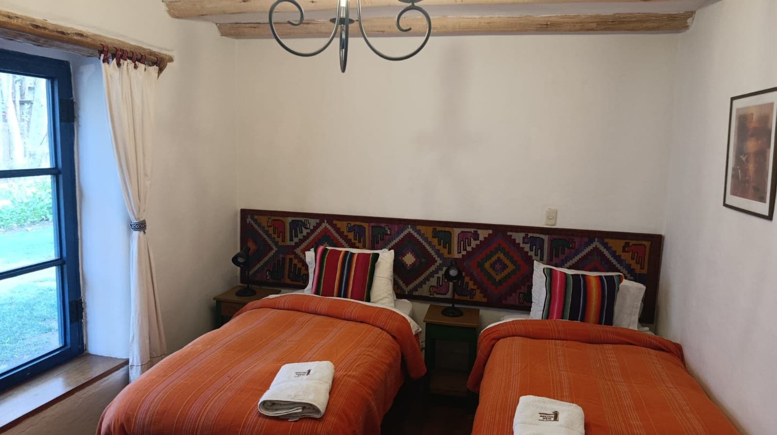 Una stanza con due letti, una finestra con tende, un muro con una testiera a motivi decorativi e un lampadario.
